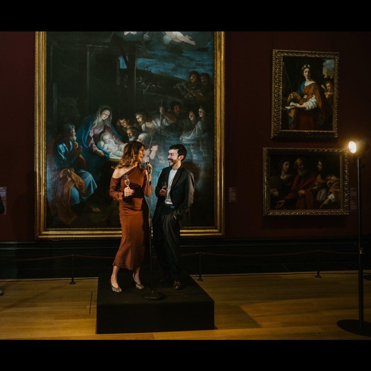 In an elegant art gallery, a woman in a rust-coloured dress and a man in a tailored jacket stand close together on a wooden platform, holding glasses of wine. Behind them, grand historical paintings adorn the deep red walls, adding a rich artistic ambiance.