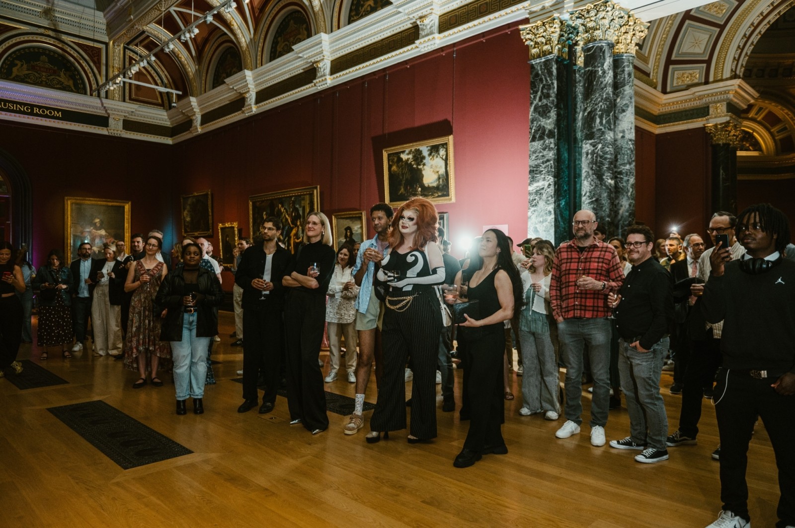 An elegant gallery setting bustling with an audience dressed in stylish attire, captivated by a performance. The room is adorned with ornate architecture, rich crimson walls, and a grand ceiling, while soft lighting illuminates the artwork and enhances the refined atmosphere.