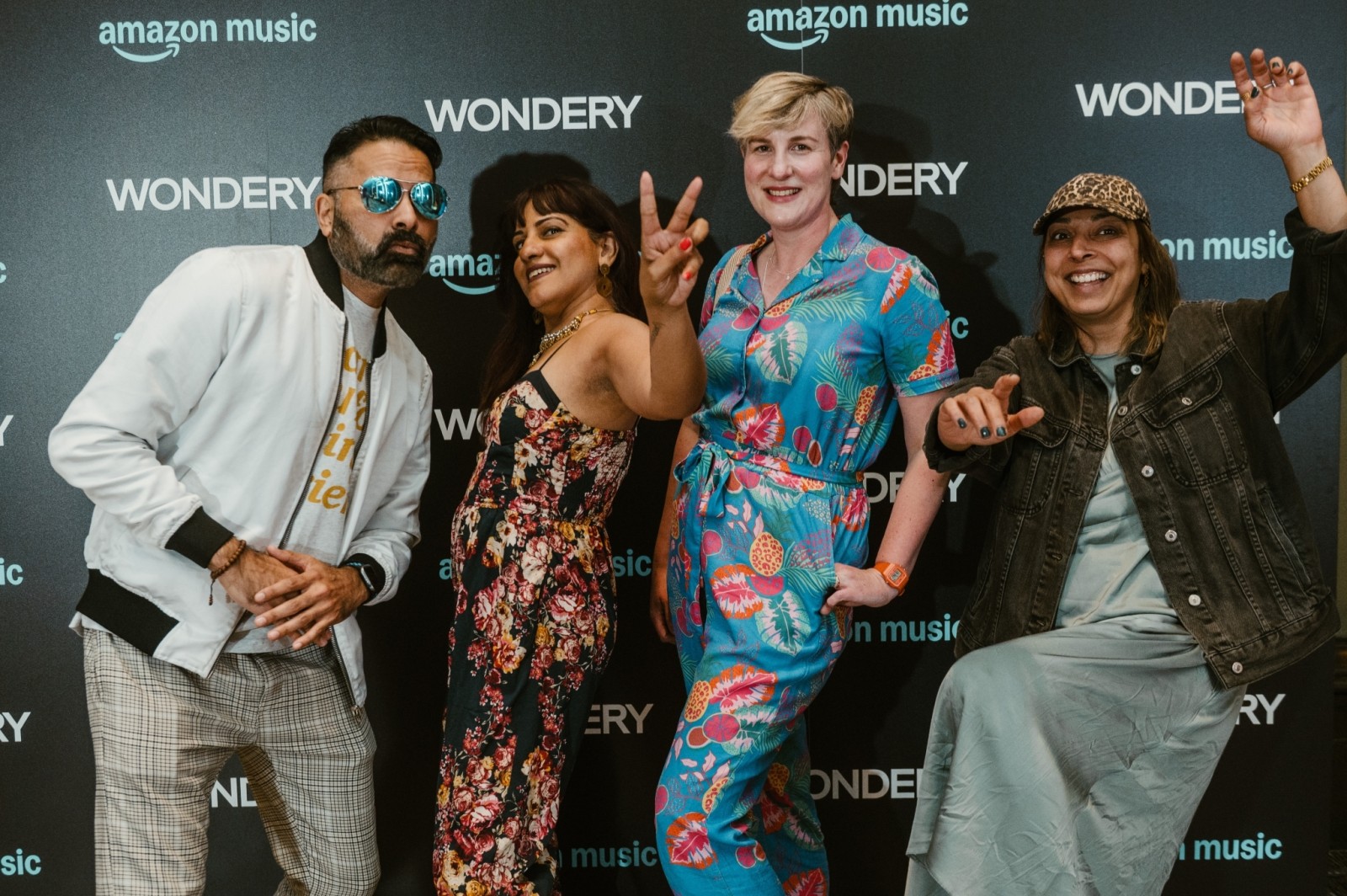 A lively group of four individuals poses playfully against a dark backdrop featuring the Wondery and Amazon Music logos. They showcase vibrant outfits: one in a bright floral jumpsuit, another in a patterned dress, complemented by casual jackets and lively hand gestures, creating an atmosphere of celebration.