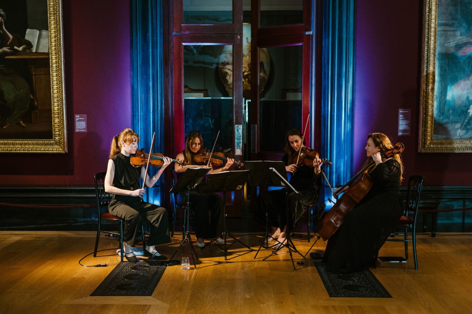A quartet of young women elegantly dressed in dark attire, each poised with their instruments—a violin, violas, and a cello—set against a rich burgundy wall. Soft purple lighting casts a serene ambience, illuminating their focused expressions as they perform in a gallery filled with classical artworks.