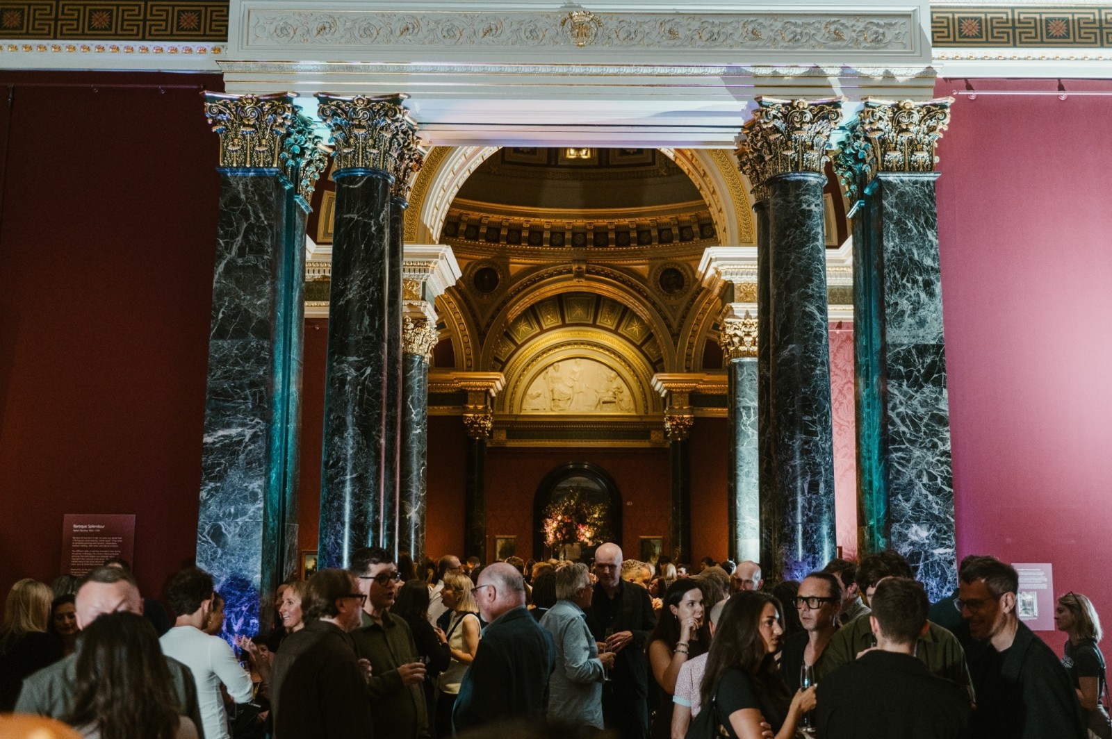A bustling gallery space adorned with grand, marble columns supporting a richly decorated archway. Guests mingle under soft lighting, creating a lively yet elegant atmosphere. The deep red walls contrast beautifully with the architectural details, enhancing the festive ambience.