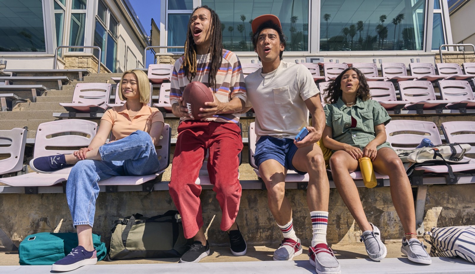 A lively group of four friends sits on bleachers illuminated by bright sunlight. The woman on the left sports a casual white T-shirt and light denim jeans, while another with long hair wears a colourful plaid shirt. The central figure holds a football, dressed in a light top and red trousers, showing a spirited enthusiasm. Next to him, another friend in a green shirt and short denim shorts enjoys a drink. The setting is vibrant, reflecting youthful camaraderie and outdoor fun.