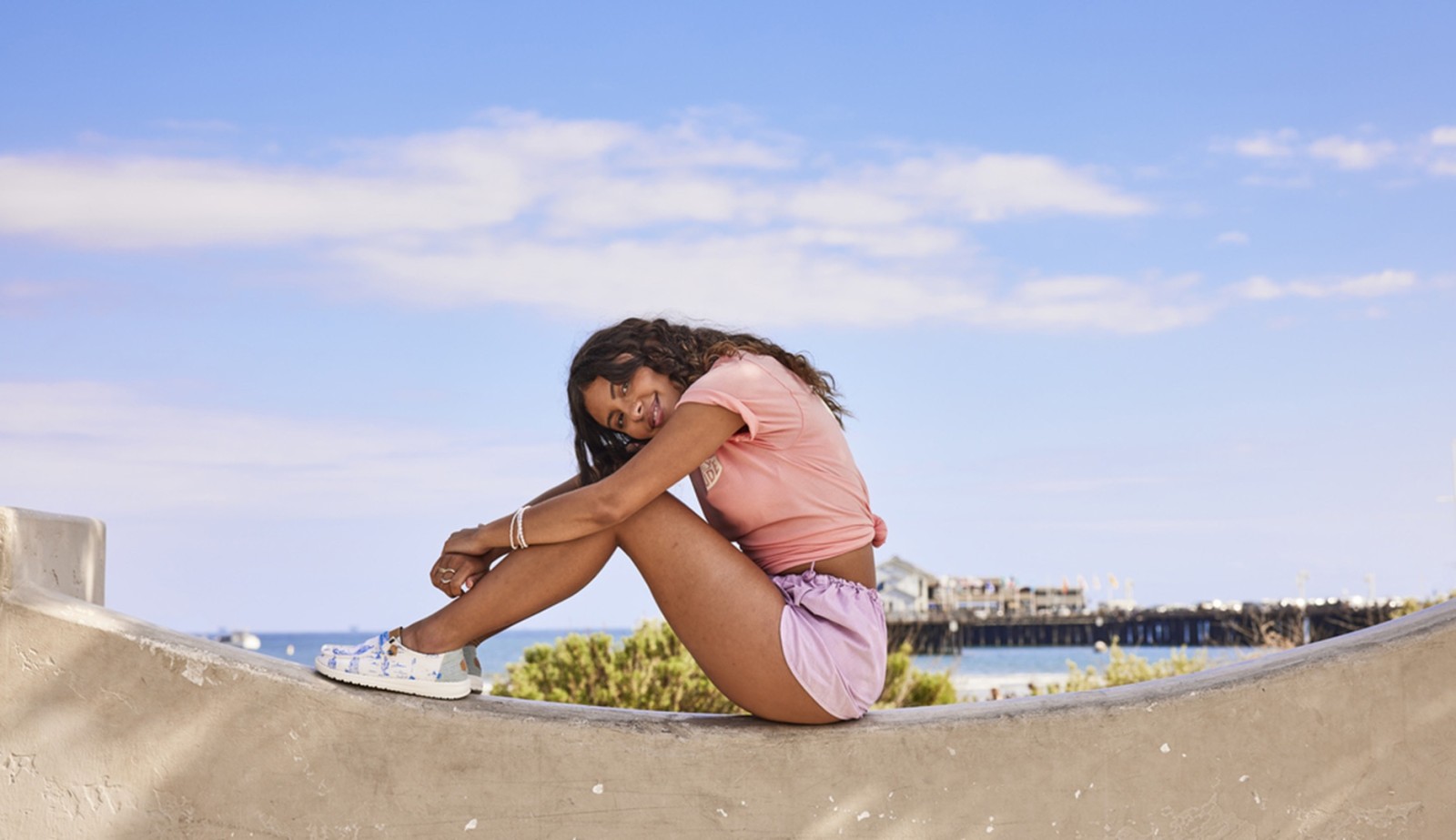 A young woman sits comfortably on a curved concrete structure, her posture relaxed as she wraps her arms around her legs. She wears a soft pink t-shirt and lilac shorts, contrasting with the blue sky and distant pier. The scene is bright and cheerful, evoking a carefree summer vibe.