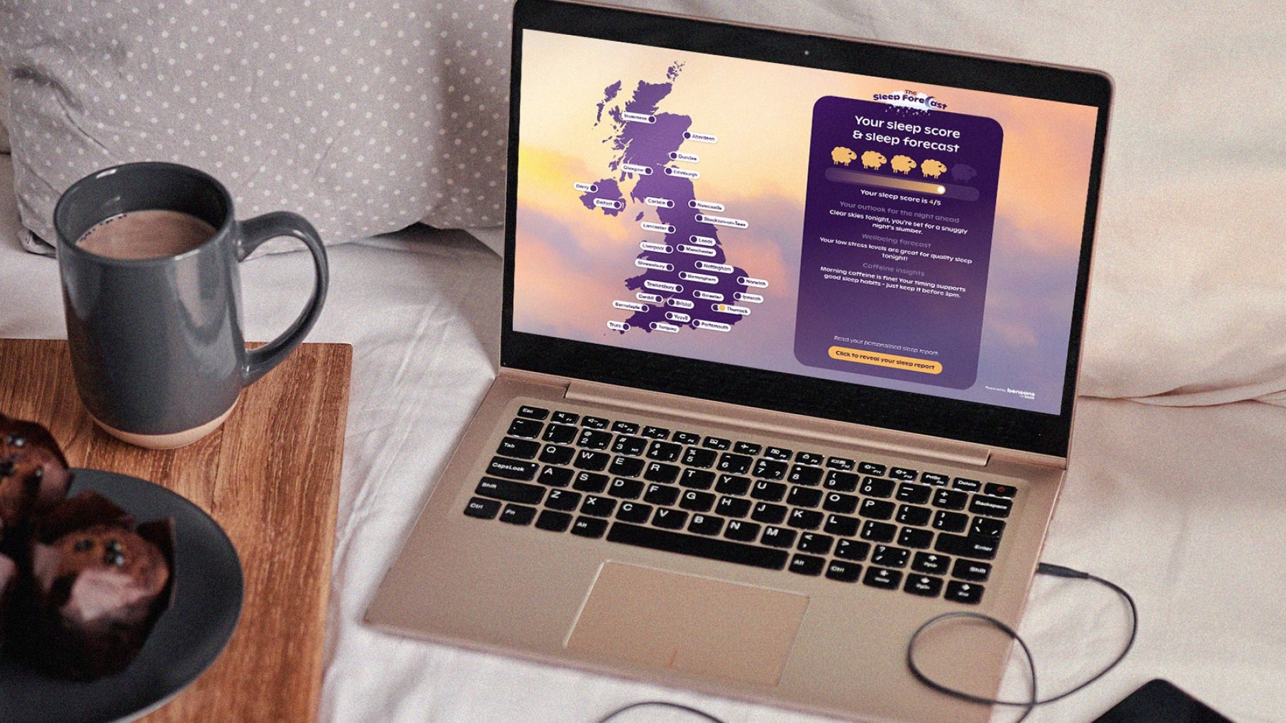 A stylish gold laptop displaying a colourful map of the UK on its screen, rests on a soft, linen blanket. Beside it, a dark grey mug filled with steaming coffee sits on a wooden tray, alongside a plate of rich chocolate muffins, invitingly decadent.
