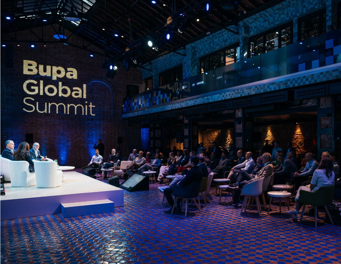 A spacious venue with high wooden ceilings features a panel discussion at the Bupa Global Summit. Three speakers sit on a white stage surrounded by an audience seated on plush chairs, illuminated by blue and amber lighting, creating a vibrant yet sophisticated atmosphere. The brick walls and mosaic flooring add character and warmth to the setting.