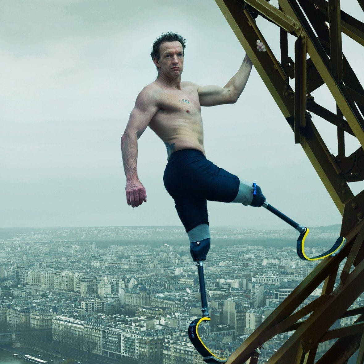 A determined man with two prosthetic legs, wearing only shorts, scales the iron framework of the Eiffel Tower. The Paris skyline stretches out beneath him, creating a striking contrast with the cloudy, overcast sky overhead, evoking a sense of adventure.