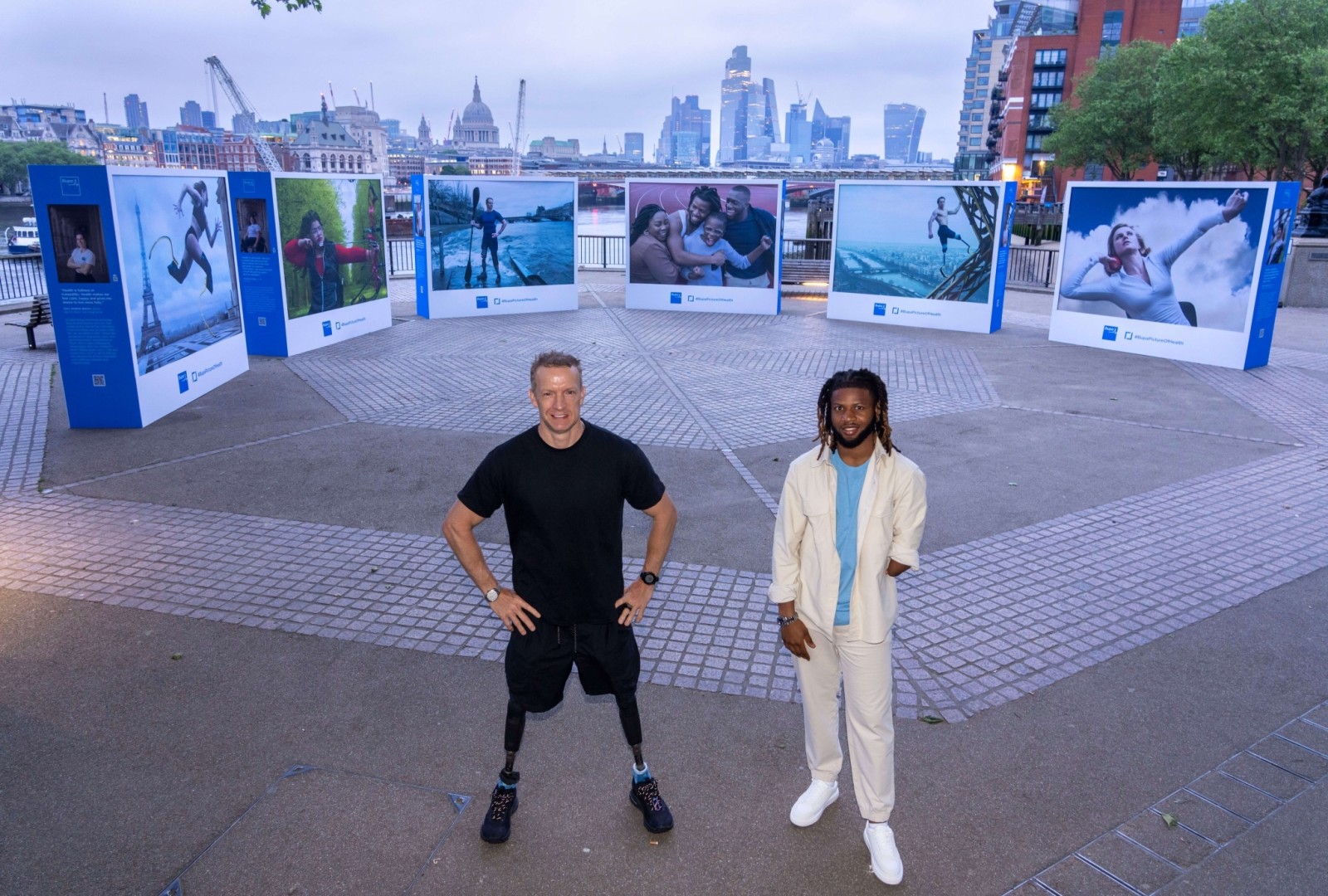 In an urban park with a view of London's skyline, two men stand confidently in front of an outdoor exhibition. One man wears a black athletic outfit, showcasing a prosthetic leg, while the other, dressed in a cream tracksuit, has long hair. Surrounding them, vibrant images depict active moments, framed elegantly in blue installations, adding a dynamic touch to the scene.