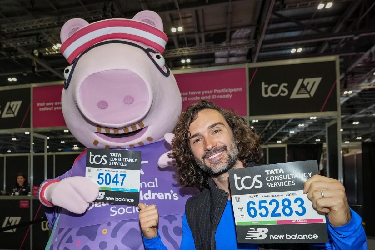 A cheerful man with curly dark hair poses with a large, friendly pig mascot wearing a red and white striped headband. Both hold race bibs featuring numbers in vibrant colors. The background showcases a bustling event area with banners highlighting Tata Consultancy Services.