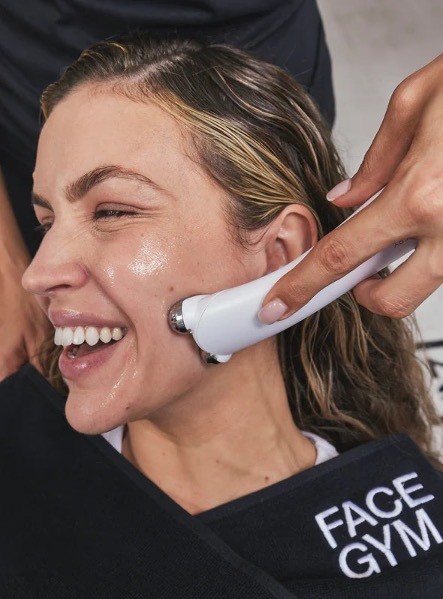 A woman with wet, wavy hair smiles brightly as she enjoys a facial treatment. A therapist's hand glides a sleek, white device along her cheek, enhancing glow with its gentle metallic surface. She is draped in a black robe featuring the logo "FACE GYM," adding a touch of elegance to the serene setting.