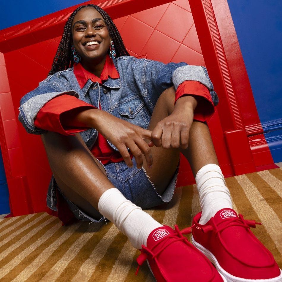A woman with braided hair sits on a striped rug, wearing a light denim jacket layered over a red shirt. Her red shoes, complemented by white ankle socks, add a pop of colour. The background features a bold red wall, enhancing the vibrant atmosphere.