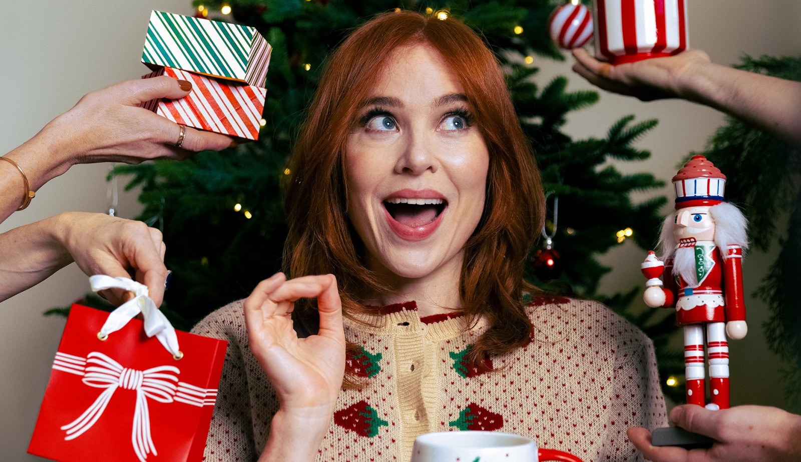 A young woman with flowing auburn hair is joyfully surrounded by festive gift boxes in vibrant red and white stripes. She wears a cozy cream sweater adorned with Christmas motifs. To her left, a decorative nutcracker and a cheerful mug complete the holiday scene, illuminated by twinkling lights from a lush green Christmas tree.