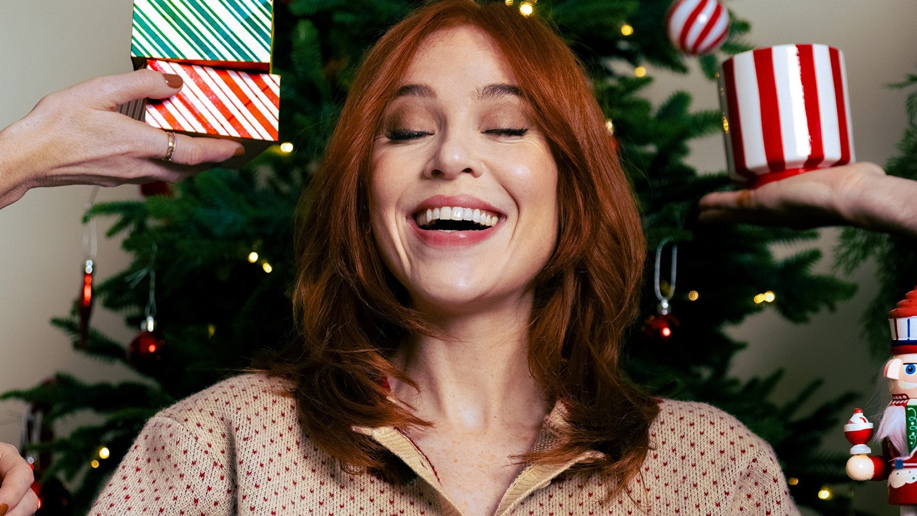A woman with chestnut hair, dressed in a festive light-coloured top, beams with joy as hands present vibrant holiday gifts adorned in red and green stripes. In the background, a decorated Christmas tree glistens with twinkling lights and shiny ornaments, creating a cheerful, festive atmosphere.