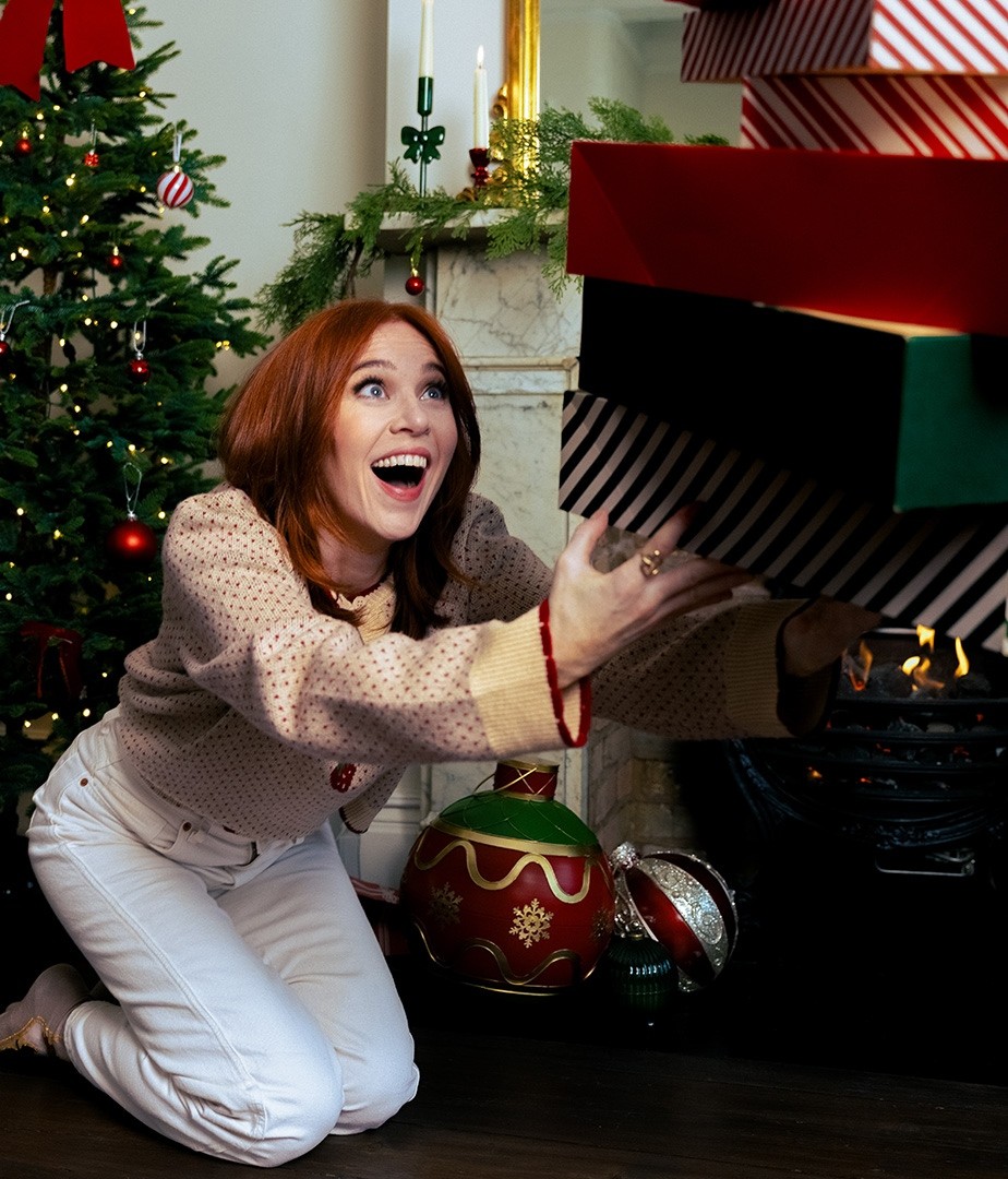 A young woman with vibrant red hair crouches on the floor, eagerly reaching towards brightly wrapped presents stacked above her. The setting features a decorated Christmas tree adorned with ornaments and twinkling lights, while a glowing fireplace adds warmth to the festive atmosphere, complemented by a colourful bauble nearby.
