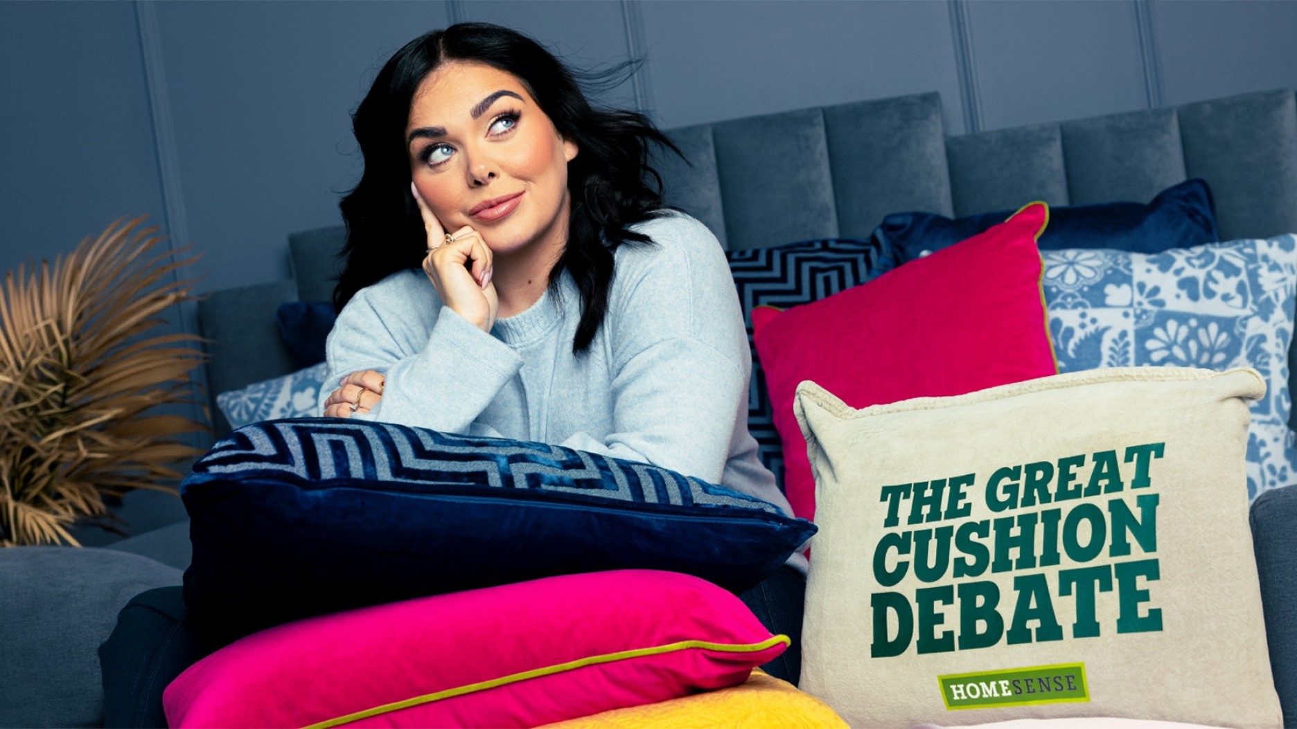 A woman with long, dark hair poses thoughtfully on a grey sofa piled high with vibrant cushions in various textures. Rich blues, bright pinks, and playful patterns create a lively contrast against the muted backdrop. One cushion prominently features the caption "THE GREAT CUSHION DEBATE" in bold green lettering, adding a whimsical touch to the scene.