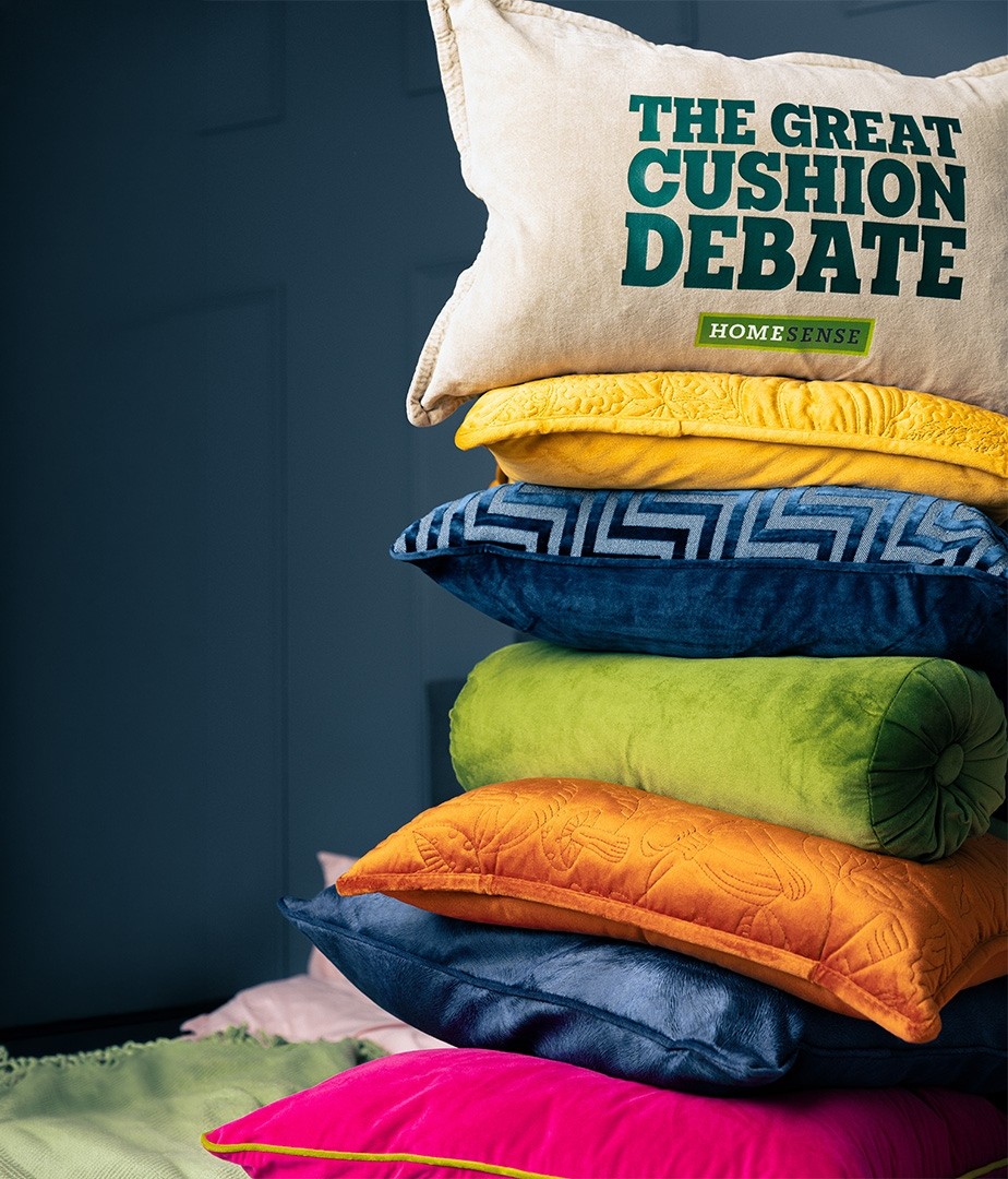 A vibrant stack of cushions, showcasing a mix of textures and colours. At the top, a cream cushion with bold green lettering reads "THE GREAT CUSHION DEBATE". Below it, rich hues of blue, green, and orange create a playful contrast, while intricate patterns add visual interest to this inviting arrangement.