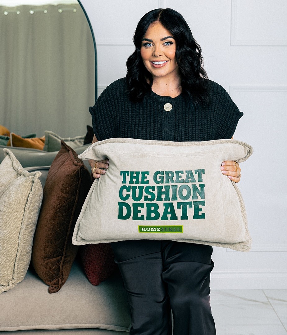 A woman with dark hair, wearing a black knitted sweater, showcases a large, cream-coloured cushion emblazoned with 'THE GREAT CUSHION DEBATE' in bold green lettering. Behind her, a stylish living space is adorned with plush, textured cushions in soft shades, creating a cozy and inviting atmosphere.