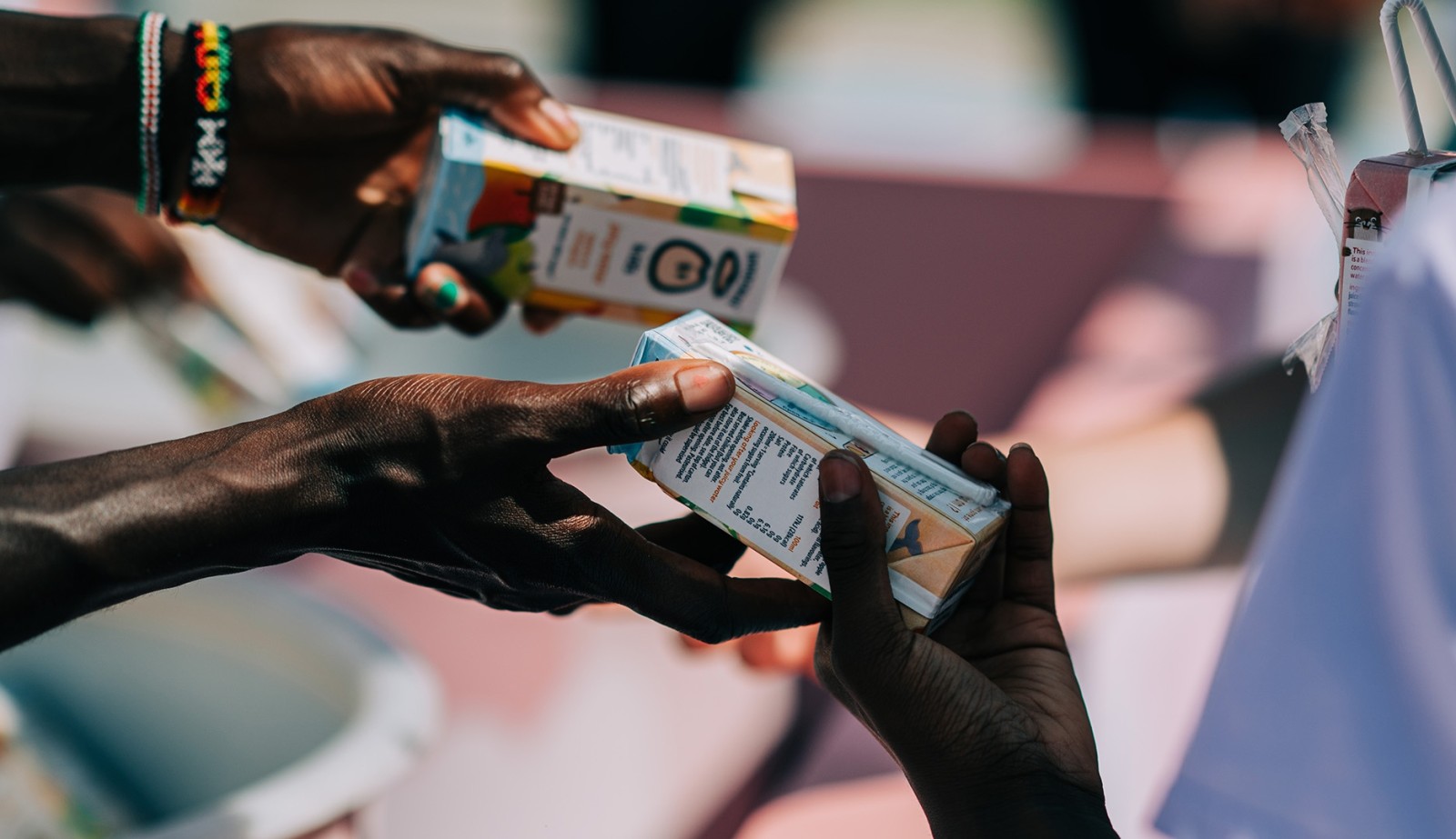 Two hands reach out to exchange colourful juice cartons under bright sunlight. The dark-skinned hand on the left holds a carton decorated with vibrant graphics, while the fair-skinned hand on the right holds another equally vibrant carton, creating a lively moment of sharing amidst a blurred background.