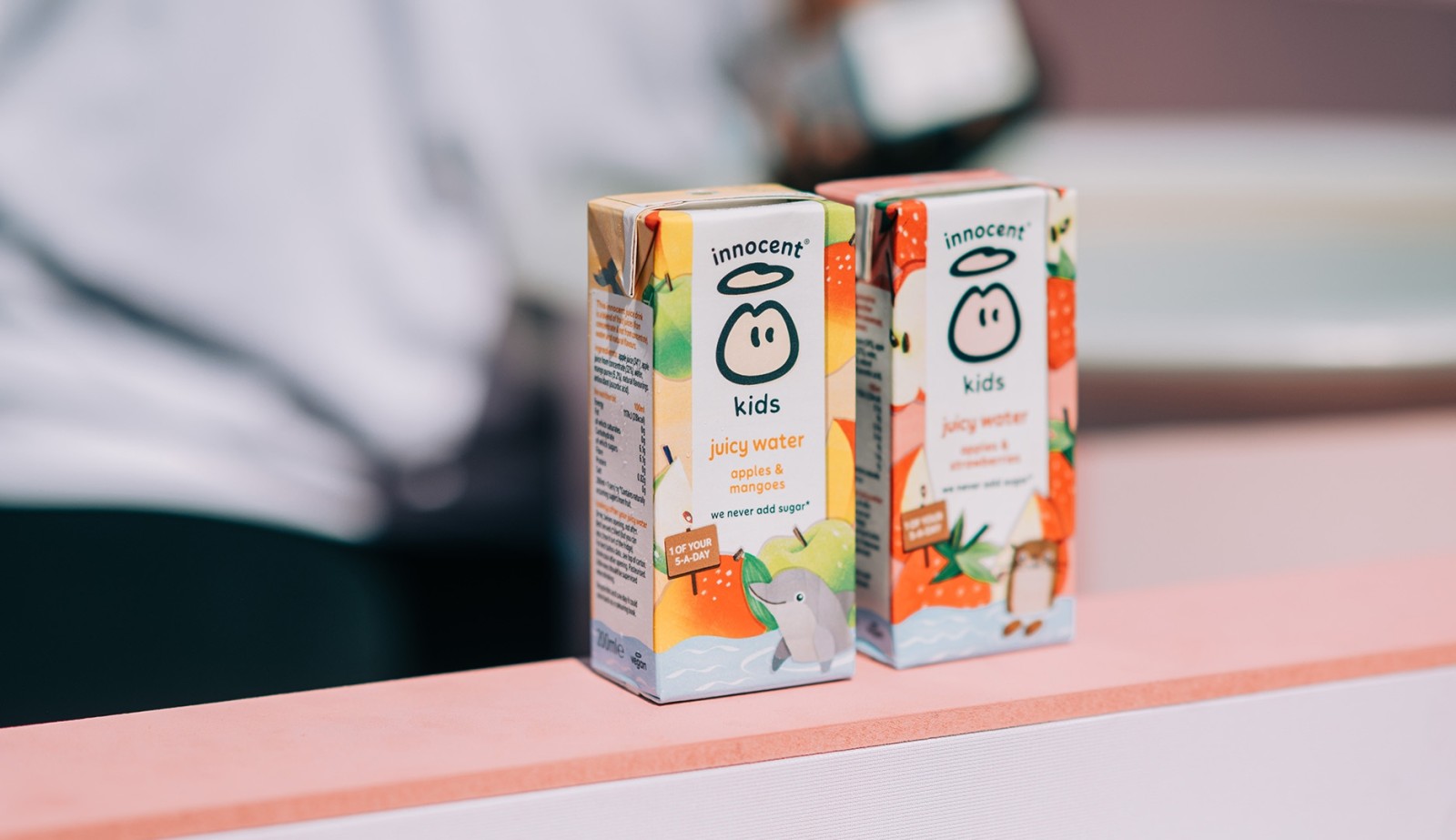 Two brightly designed cartons of Innocent Kids Juicy Water rest on a pastel pink countertop. Each carton features playful illustrations of a dolphin and tropical fruits, exuding a fun and vibrant appeal. A blurred figure in the background adds a sense of lively atmosphere.