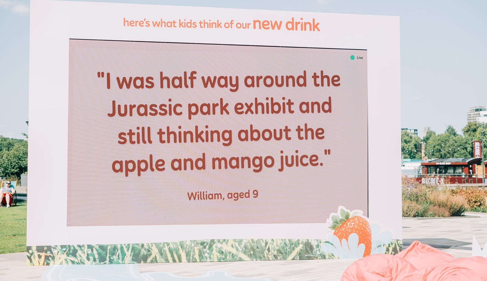 A large, light-coloured backdrop displaying a quote from a child about a drink, surrounded by vibrant green foliage illustrations at the base. The quote, in playful, oversized text, reads: "I was half way around the Jurassic park exhibit and still thinking about the apple and mango juice." below it, the name William, aged 9, is presented in a smaller font.
