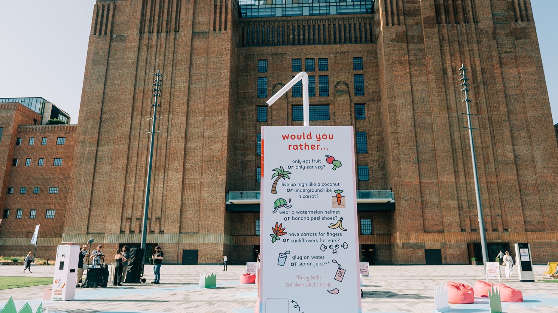 Against the backdrop of a grand, red-brick building, a large white sign with pink typography poses a whimsical question: "Would you rather…". The sign is adorned with playful illustrations of various foods and scenarios, inviting passersby to engage. The open plaza is bustling with people, and the sunlight creates a vibrant, inviting atmosphere.