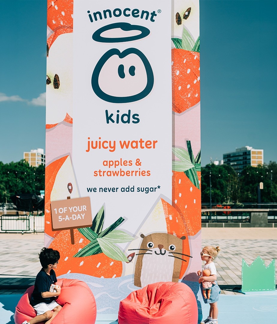 A vibrant outdoor scene featuring a large, colourful promotional banner for Innocent Kids' juicy water, adorned with playful illustrations of strawberries and cheerful creatures. In the foreground, two children play on soft, oversized pink bean bags, surrounded by lush greenery and a clear blue sky.