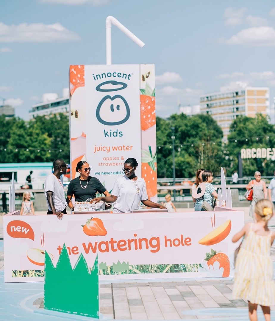 A vibrant outdoor scene featuring a large, cheerful sign for 'Innocent Kids Juicy Water' adorned with playful graphics of apples and strawberries. Two team members, dressed in branded shirts, engage with children nearby, while a sunny blue sky enhances the lively atmosphere of a playful public space.
