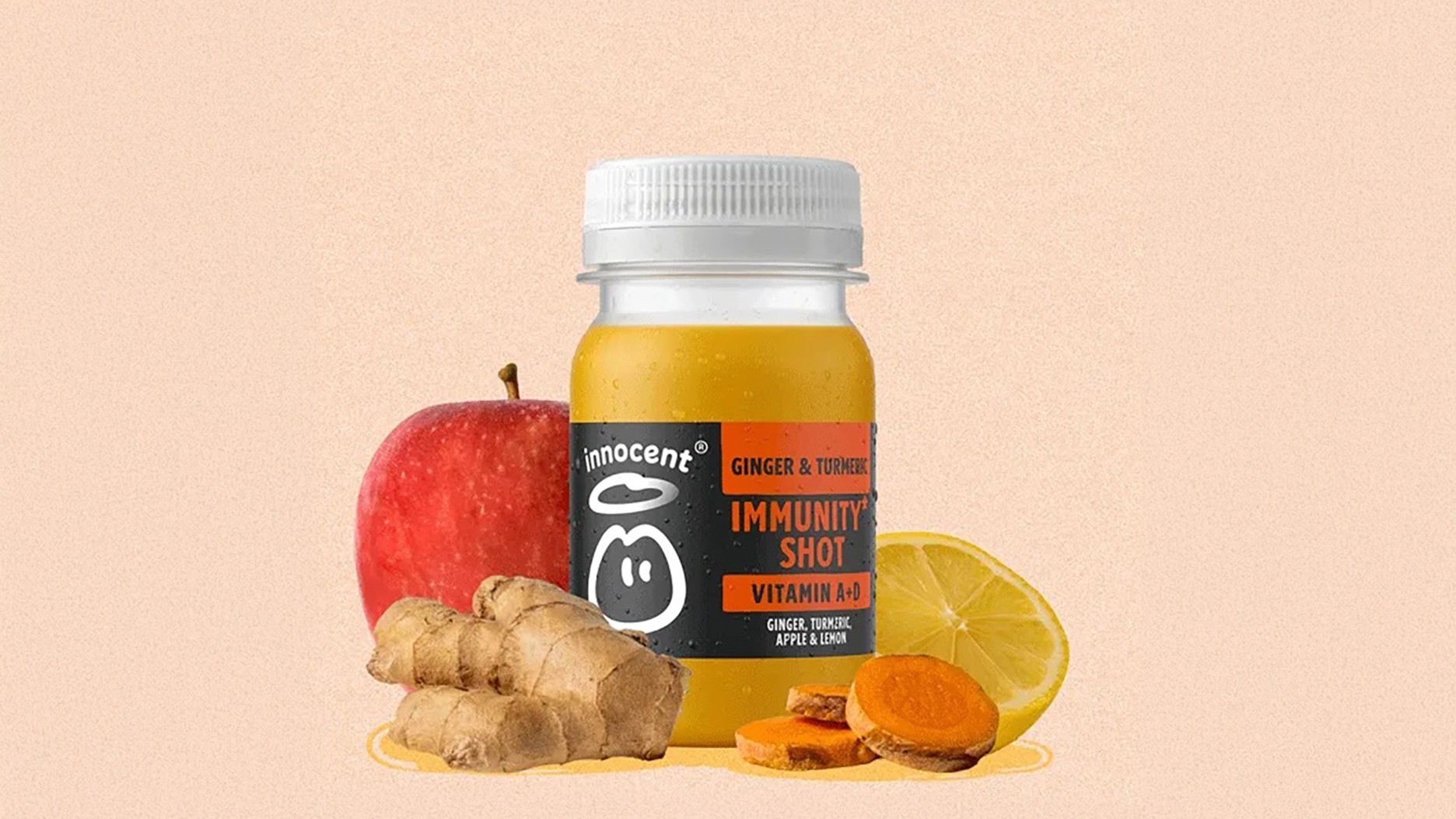 A glass jar filled with a vibrant yellow immunity shot, labelled 'Innocent' and featuring ginger, turmeric, and lemon, sits against a soft peach background. Fresh ginger root and slices of lemon are arranged beside the jar, along with a shiny red apple, showcasing a healthy, refreshing composition.