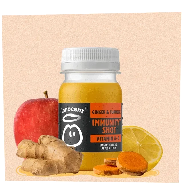 A clear glass bottle of ginger and turmeric immunity shot stands prominently against a light peach background. The vivid yellow liquid inside contrasts with the earthy ginger root in the foreground and vibrant red apple, alongside bright lemon slices. The bottle features a playful label, enhancing the fresh, health-focused aesthetic.