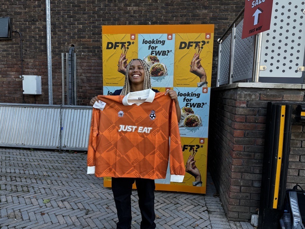 A young individual stands confidently, wearing a vibrant orange sports jersey emblazoned with "JUST EAT" on the front. They smile broadly, holding the oversized jersey. In the background, a colourful promotional poster features hands holding food, set against a brick wall, enhancing the lively atmosphere.