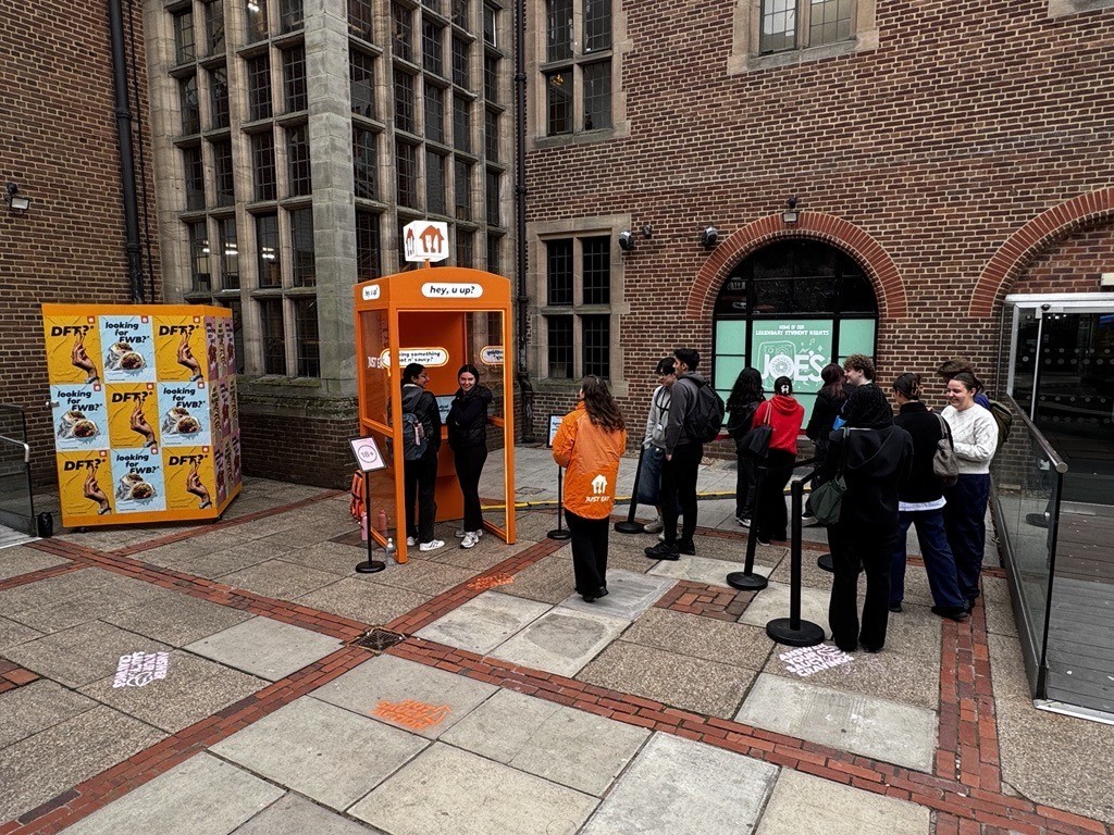 A bustling outdoor area features a vibrant orange food stall adorned with playful graphics. A queue of people, clad in coats, waits eagerly beside it, while the textured brick and stone architecture of nearby buildings frames the scene. Soft daylight illuminates the vibrant atmosphere.
