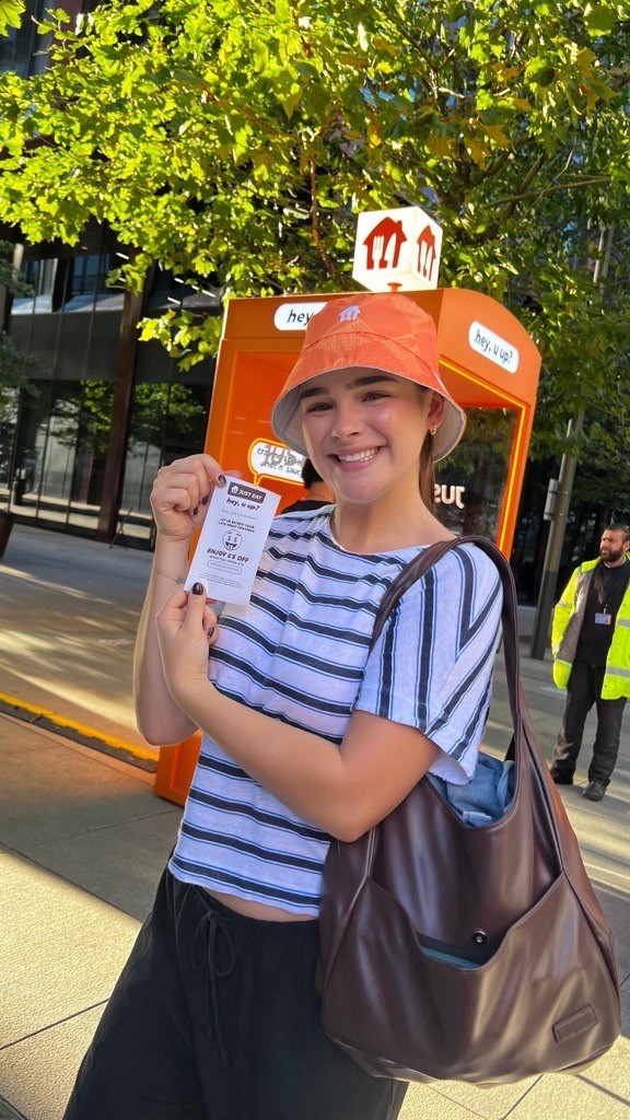 A smiling young woman stands outdoors, holding a ticket in her right hand. She wears a striped t-shirt and an orange bucket hat, complemented by a stylish brown handbag slung over her shoulder. Behind her, vibrant green trees frame a bright urban setting.