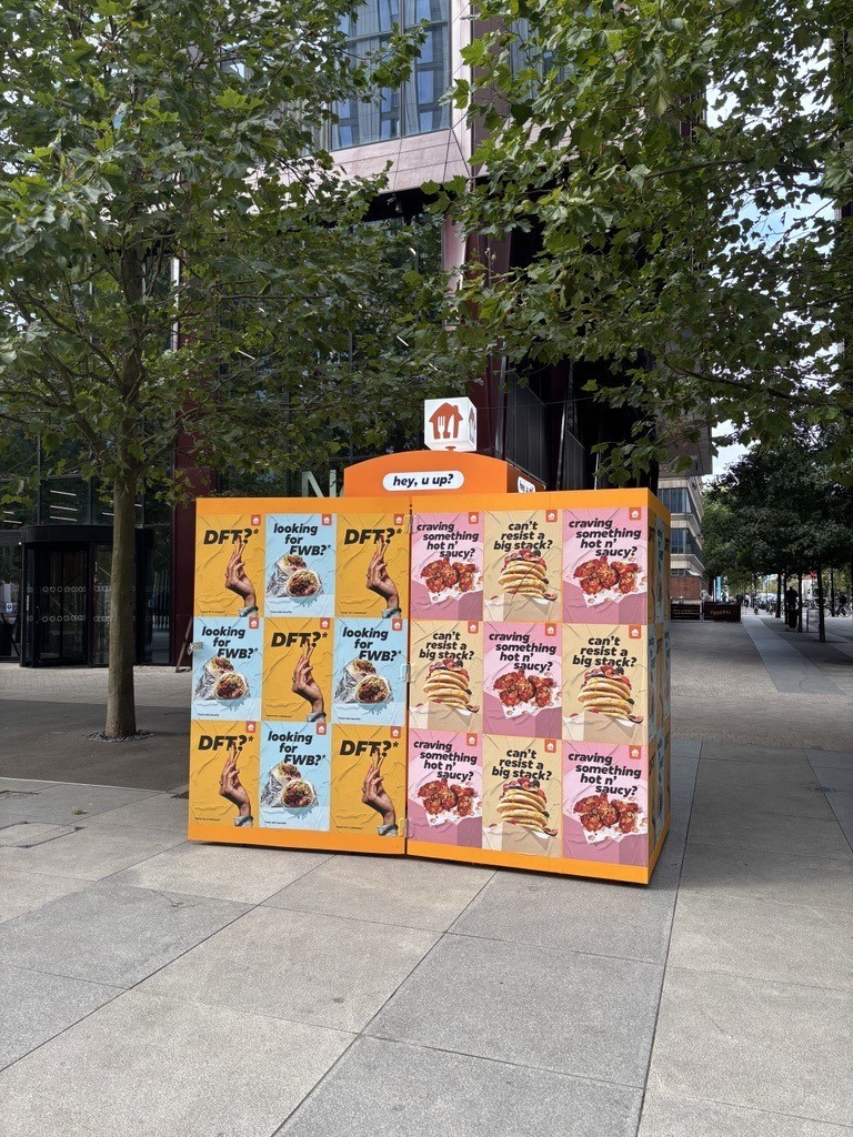 A colourful promotional kiosk stands prominently on a pavement, showcasing vibrant posters on each side. The blue and pink designs feature enticing food imagery, with playful typography, amidst leafy trees providing a lively backdrop while sunlight creates dappled shadows.