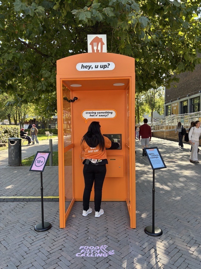 A vibrant orange telephone booth with a rounded top stands in a bustling outdoor area. A woman in an orange jacket is seen interacting with a digital screen inside. Surrounding the booth are pedestrians and trees, with signage on the ground indicating food-related themes.