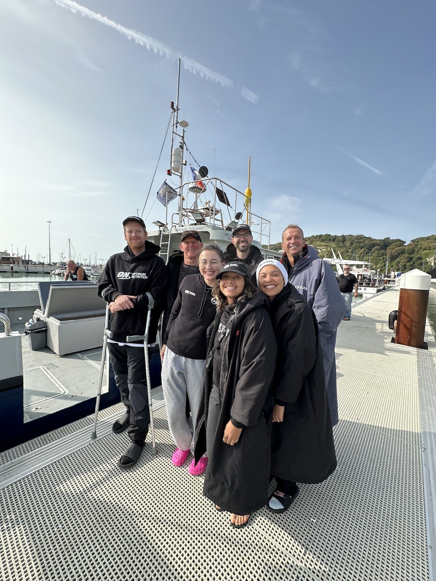 A group of seven individuals poses on a sunlit marina dock, surrounded by boats. They are dressed in casual clothing, including cosy jackets and comfortable footwear. The backdrop features a gently rippling harbour, with hills in the distance and a clear blue sky overhead.