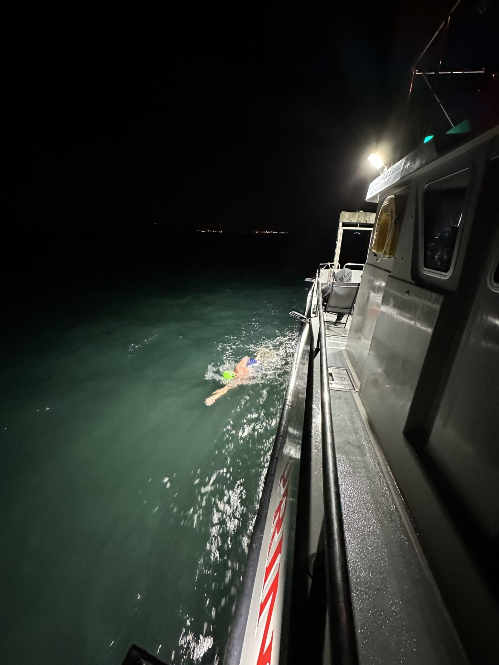A swimmer dons bright swim gear, partially illuminated by the nearby rescue boat, cutting through dark waves under a starry night. The boat’s silver hull contrasts against the deep green water, creating a striking maritime scene. The smooth surface reflects faint lights from the shore.