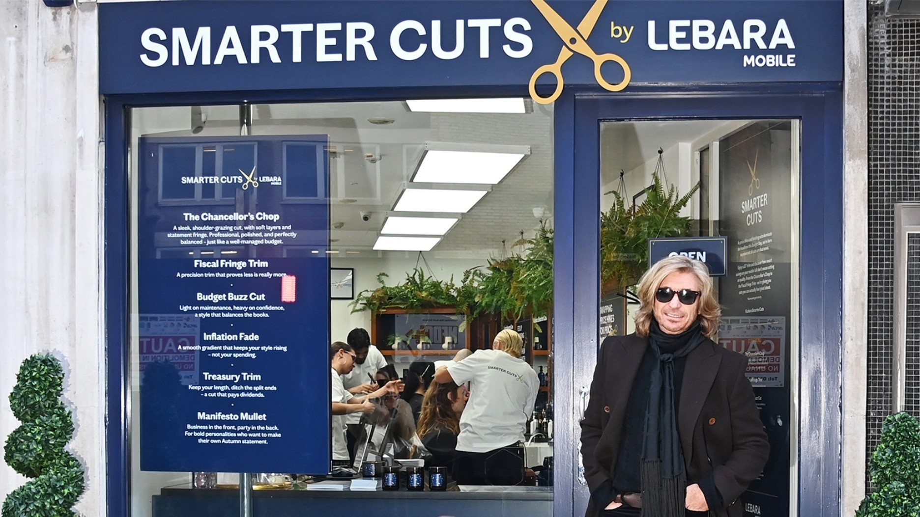 The entrance of a chic barbershop named "Smarter Cuts by Lebara Mobile" features a bright window with a modern layout showcasing a lively interior. An elegant woman with light hair stands confidently in black attire, against a backdrop of green potted plants, while customers are visible inside.