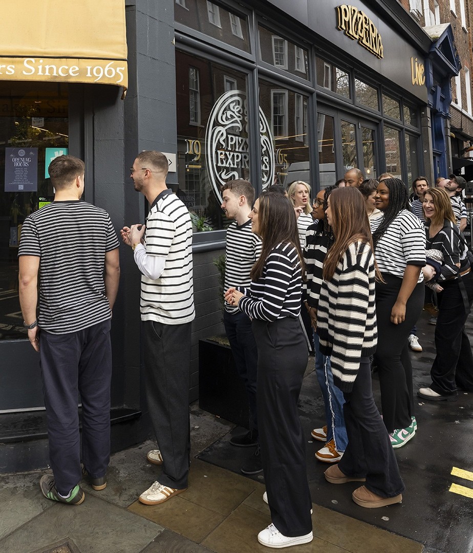 A lively group of individuals, dressed in matching black and white striped shirts, stand in a line eagerly outside a bustling pizzeria in an urban setting. The shop's warm yellow awning contrasts with the grey facade, adding a vibrant touch to the scene.
