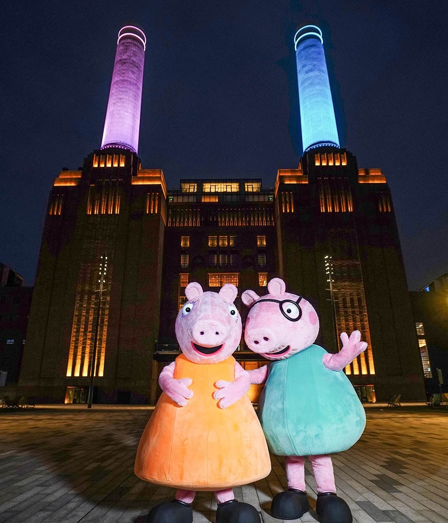 Two large, animated pig characters stand in front of the iconic Battersea Power Station, illuminated at night. The vibrant pinks and blues of the characters contrast with the striking backdrop of the historic, industrial architecture, beautifully lit with multicoloured lights.