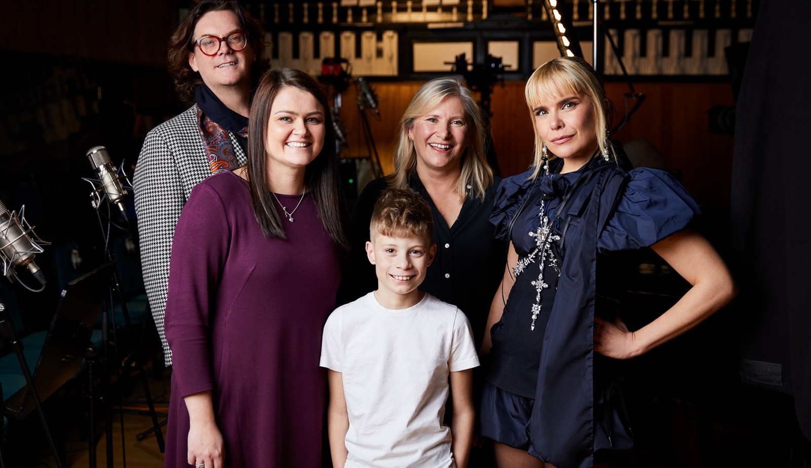 A lively group of five stands in a recording studio, surrounded by microphones and sound equipment. The women are dressed stylishly, with one in a chic black outfit and another in a deep purple dress, while a young boy in a white t-shirt stands among them, smiling brightly. Exotic wood paneling and musical notations form the backdrop, enhancing the creative atmosphere.