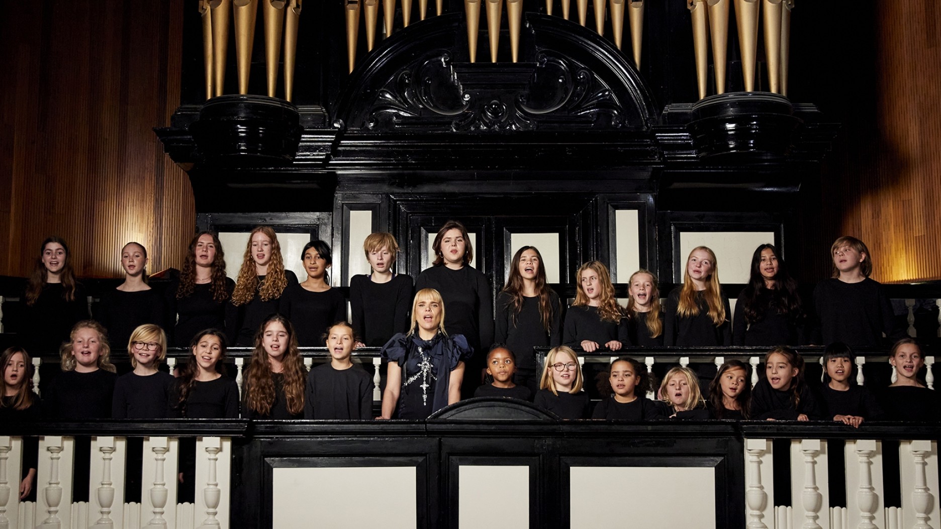 A large choir of children stands on a grand balcony, dressed in simple black attire. They are arranged in rows, with the backdrop of intricate wood paneling and elegant organ pipes rising above them, creating a contrast of rich textures and warm tones in the illuminated space.