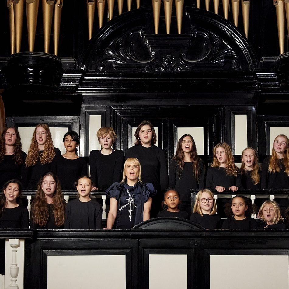 A diverse group of young singers stands on a wooden balcony, clad in black attire that contrasts with the dark, ornate woodwork around them. The choir members are positioned in a semicircle, harmonising under the grandeur of a vintage organ, its golden pipes reaching skyward. Delicate light spills into the space, highlighting the textures of their outfits and the intricate design of the architectural elements.