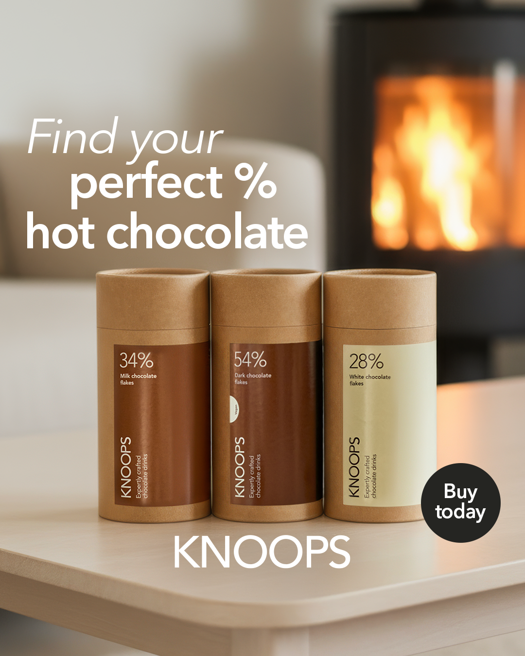 Three stylish cylindrical containers of KNOOPS hot chocolate sit on a light wooden table. The containers are crafted from eco-friendly brown and cream paper, labelled with elegant percentages: 34%, 54%, and 28%. A warm, glowing fireplace flickers softly in the blurred background.