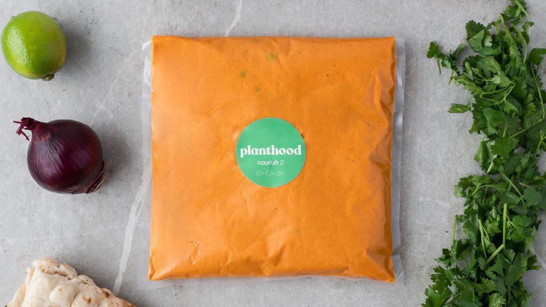 A vibrant orange sauce packet with a circular green label reading "planthood" sits at the centre of a light grey marble countertop. Surrounding it are fresh ingredients: ripe red cherry tomatoes, a zesty lime, a red onion, and fragrant cilantro, alongside a piece of toasted flatbread, adding a rustic touch to the composition.