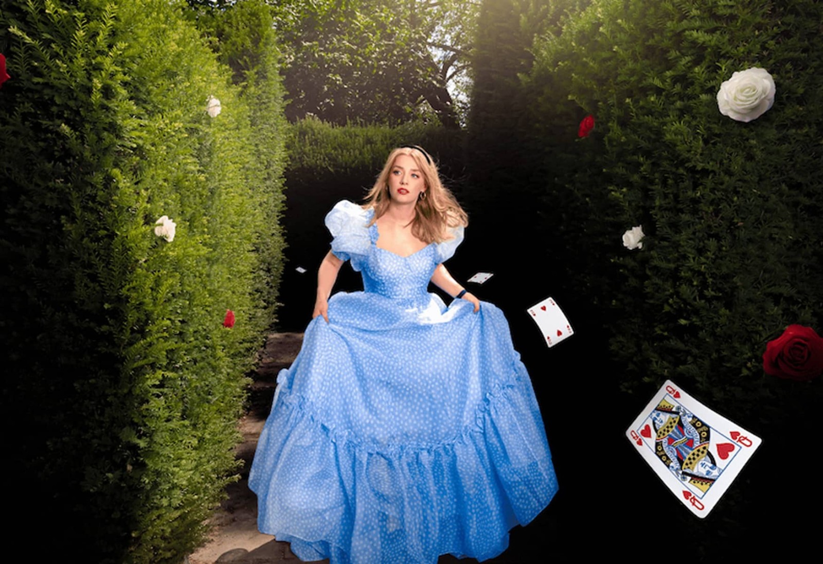 A young woman wearing a flowing, powder blue gown adorned with white polka dots navigates a lush hedge maze. The sunlight dapples through the dense greenery, while playing cards and vibrant red roses float whimsically around her, enhancing the enchanting atmosphere.