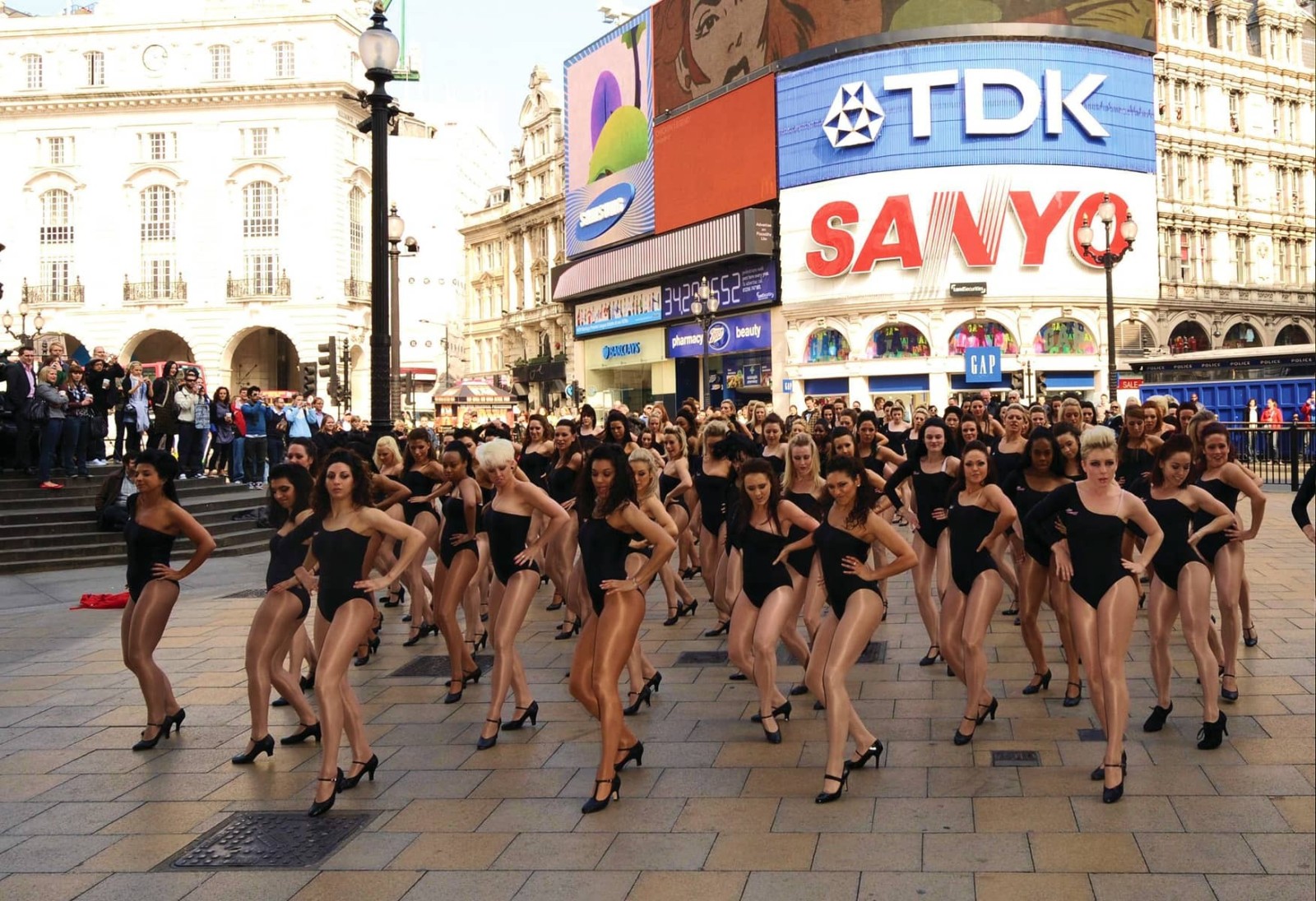 A vibrant scene in a bustling square showcases a large group of dancers, all clad in sleek black bodysuits and high heels, gracefully performing a choreographed routine. The sunlight enhances the polished surfaces and creates a lively atmosphere, while iconic advertisements frame the backdrop.