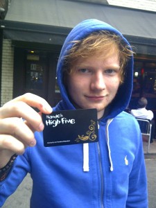 A young man with tousled ginger hair holds up a black loyalty card labelled "Nando’s High Five," adorned with ornate golden swirls. He wears a bright blue zip-up hoodie, standing in front of a casual restaurant setting. Soft afternoon light filters through the location, creating a relaxed vibe.