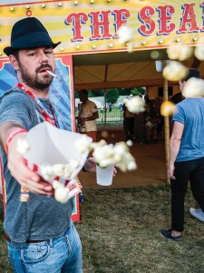 A man wearing a black fedora and a grey t-shirt gestures towards the viewer, offering popcorn cascading from a crisp white cone. He stands in front of a vibrant circus-themed stall, while festival-goers mingle in the background, illuminated by soft evening light.