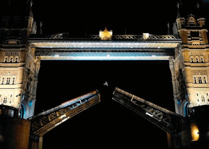 The iconic Tower Bridge stands majestically against the dark night sky, illuminated by soft blue and golden lights. At its centre, the bridge's two towers rise impressively, while the road sections on either side are raised, creating an elegant arch. This dynamic structure showcases its intricate Victorian design, framed by the gentle reflections on the River Thames below.