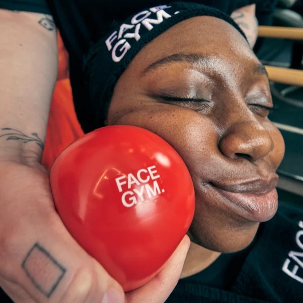 A close-up of a young woman with smooth skin relaxing as her eyes are closed, while a hand gently presses a bright red, round ‘FACE GYM’ ball against her cheek. The scene is set in a stylish gym with natural light creating a soft ambience, enhancing the vibrant colours.