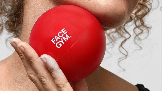 A close-up of a woman holding a vibrant red facial massage ball against her jawline, showcasing its smooth, glossy surface. The ball features the logo "FACE GYM" in bold white lettering. Curly hair frames her face, highlighting the minimalist aesthetic of her black top and the simplicity of the background.